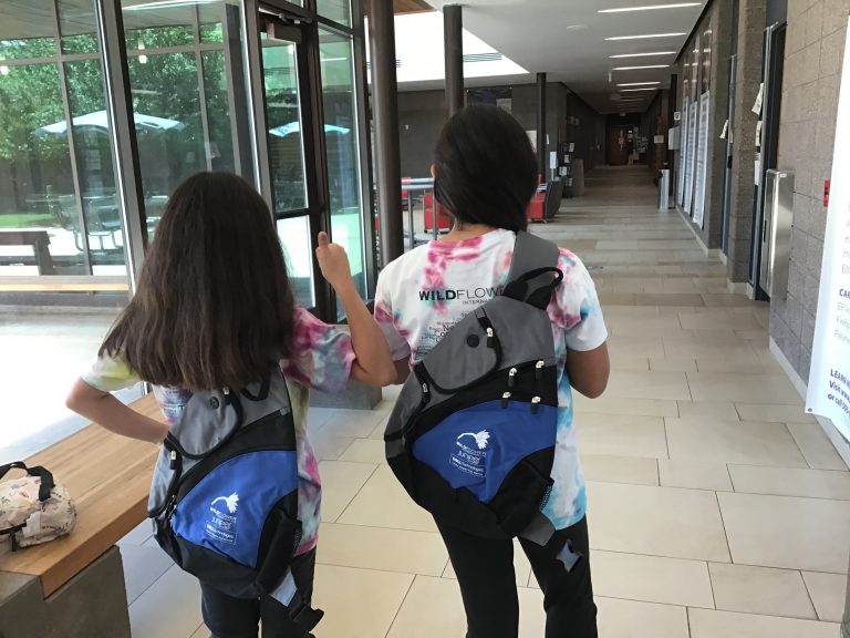 Two students with backpacks and Wildflower t-shirts walking in a school hallway.