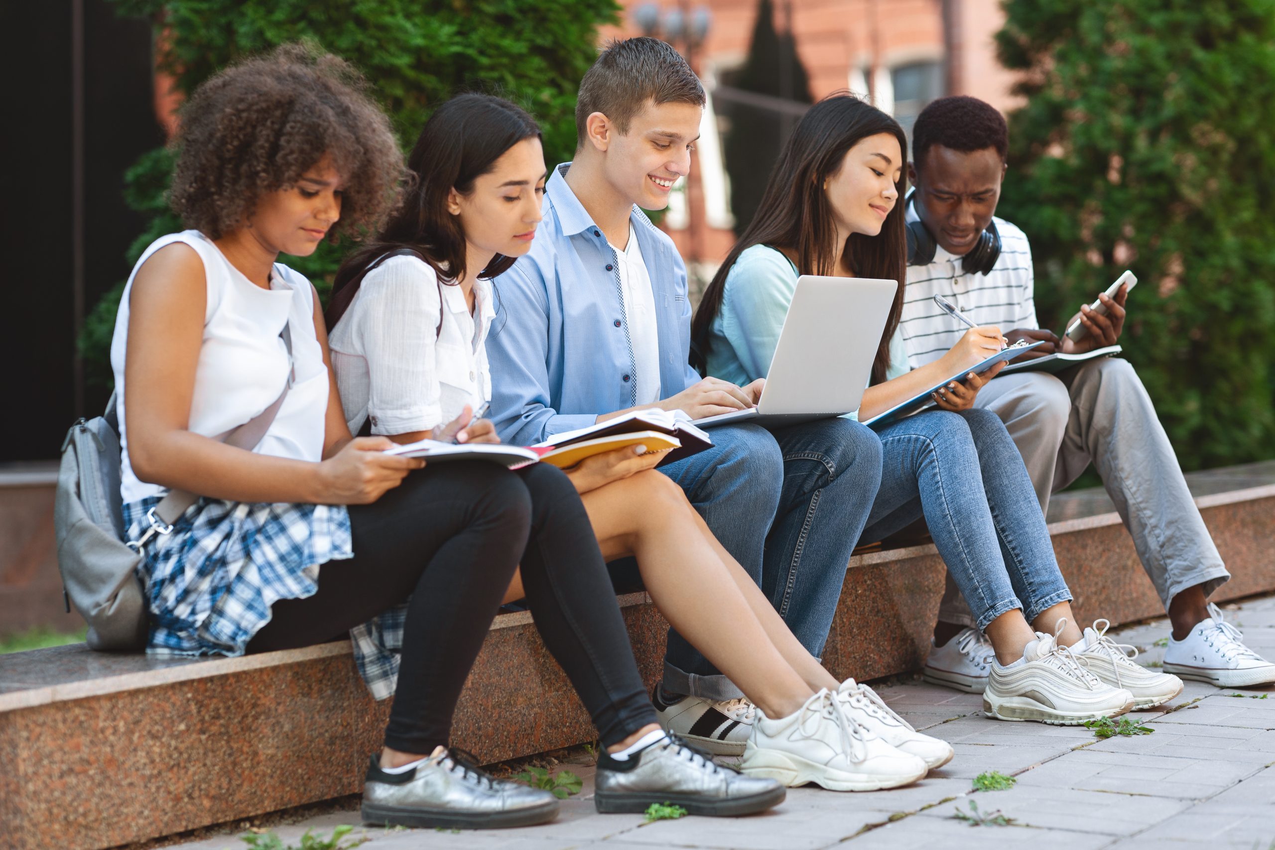 A group of teens reading and taking notes seated on a bench.