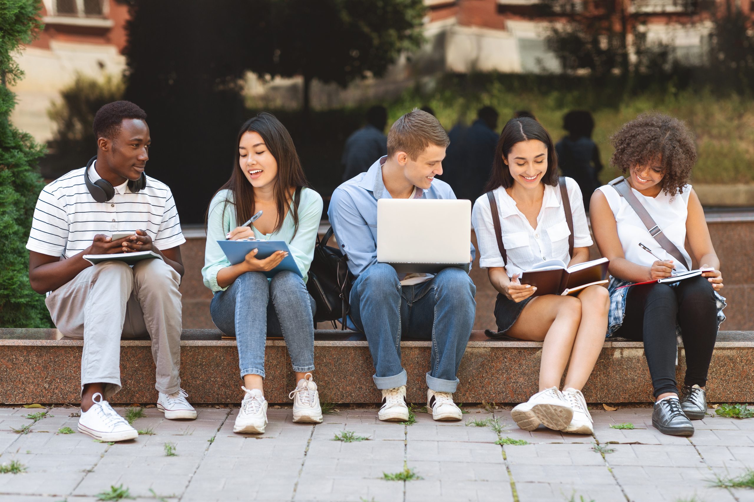 A group of teens seated on a curb, discussing with open books and laptops.